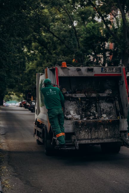 A group of five individuals dressed in light blue T-shirts and casual trousers participating in a woodland cleanup activity. They are collecting trash in a forested area with tall trees and dappled su