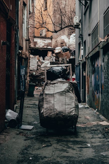 A narrow, urban alleyway cluttered with overflowing rubbish bags and loose trash scattered across the asphalt surface. In the foreground, a large, weathered grey plastic refuse sack with visible creas