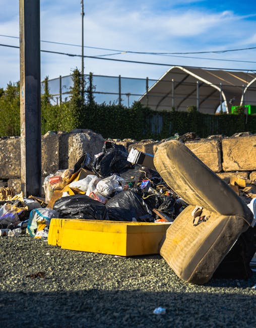 A narrow, urban alleyway cluttered with overflowing rubbish bags and loose trash scattered across the asphalt surface. In the foreground, a large, weathered grey plastic refuse sack with visible creas