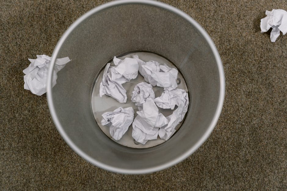A cylindrical waste bin with a metallic mesh exterior filled with crumpled white papers is positioned on a carpeted floor. The metal finish of the bin has a matte, slightly textured surface with a gre