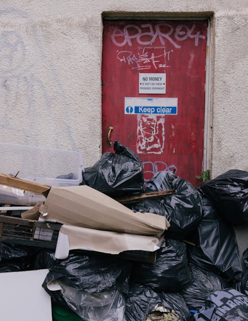 A large, green metal skip filled with various waste materials and cardboard packaging, situated outdoors on a paved area. The contents include flattened cardboard boxes, some partially collapsed, with